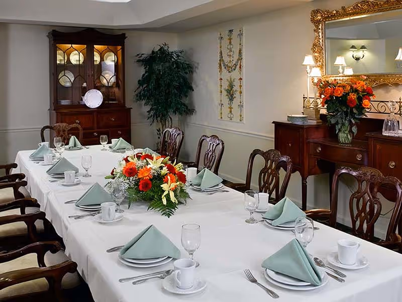 A formal dining room with a long table covered with a white tablecloth, set with plates, cups, glasses, silverware, and green folded napkins. A large floral centerpiece with orange and white flowers is in the middle of the table. The room features wooden chairs with cushioned seats, a wooden cabinet with glass doors displaying plates, a sideboard with a large ornate gold-framed mirror above it, and a vase of orange flowers. A potted plant and decorative wall art are visible in the background.