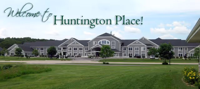 Front exterior view of the Huntington Place senior living building with a large lawn and the words 'Welcome to Huntington Place!' across the sky.