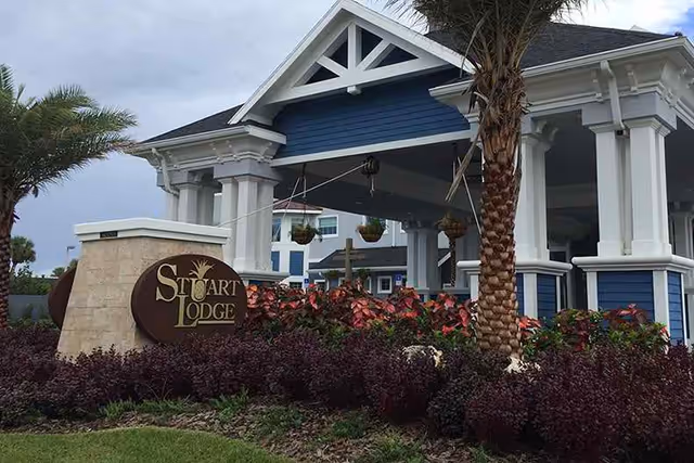 Entrance of a blue-and-white building with a porte-cochere, a 'Stuart Lodge' sign, palm trees, and landscaped plants.