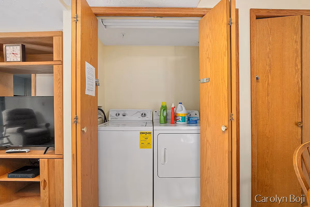 Washer and dryer inside an open wooden closet with laundry supplies on top, next to a TV cabinet and chair.