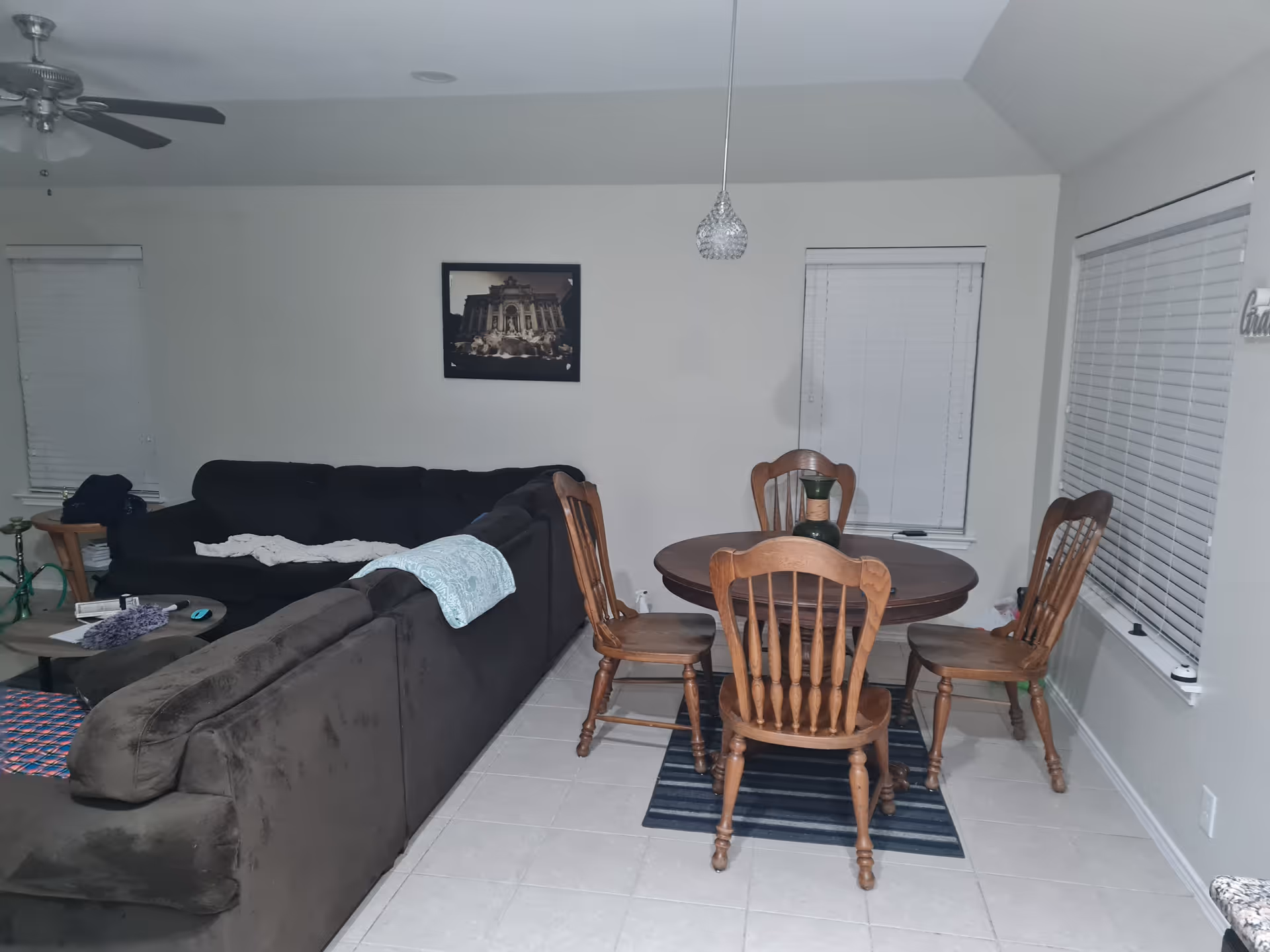 Interior view of a living and dining area with a dark sectional sofa, a round wooden dining table with four wooden chairs, two windows with blinds, a ceiling fan, a hanging light fixture, and a framed picture on the wall.