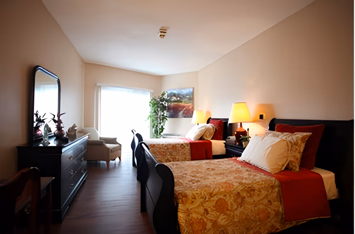 A warmly lit bedroom with two single beds, each with patterned bedspreads and red accents. There are two bedside tables with lamps, a large window with sheer curtains, a dresser with a mirror, and a comfortable armchair near the window. A plant and a framed picture are visible in the corner.