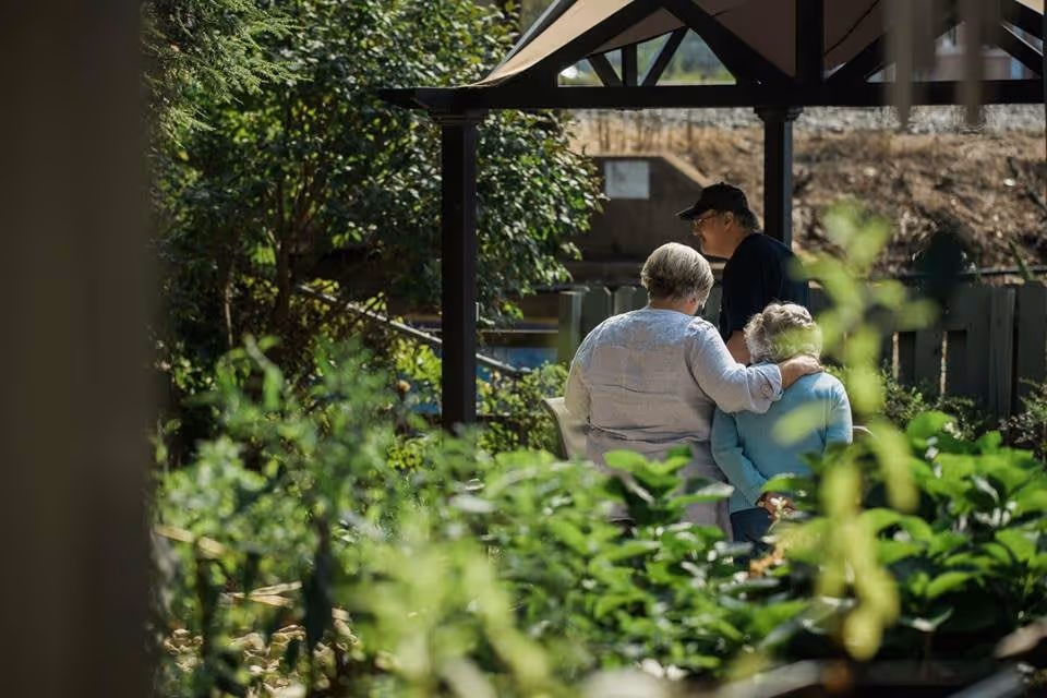 Three elderly people sitting and standing together in a garden area with green plants and trees around them. One person is sitting with their arm around another, while a third person stands nearby under a pergola structure.