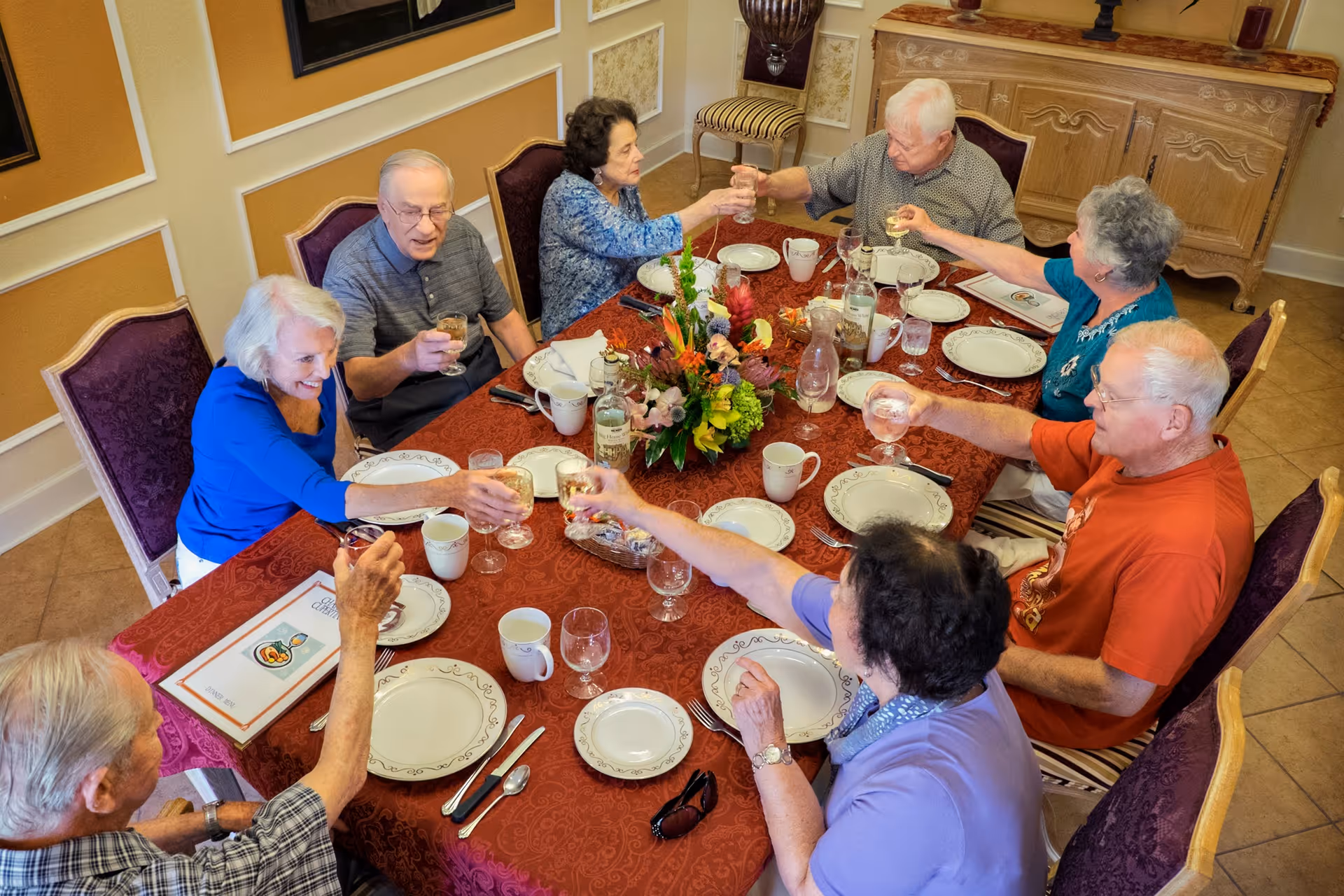 A group of elderly people sitting around a dining table with a red tablecloth, raising their glasses in a toast. The table is set with plates, cups, utensils, and a floral centerpiece. The room has beige walls with white trim and a wooden sideboard in the background.
