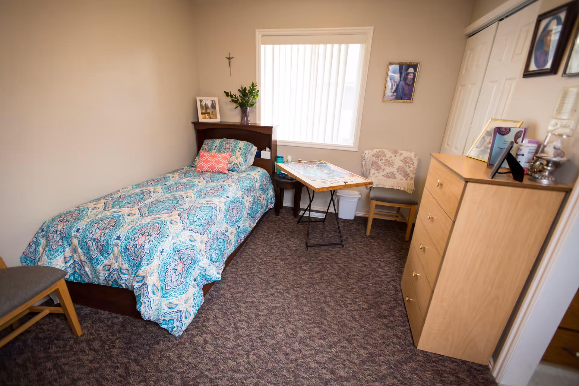 A small, cozy bedroom with a single bed covered in a blue and white patterned bedspread. There is a wooden headboard with a framed photo and a vase with green leaves on it. A folding table with a puzzle on it is placed near a window with vertical blinds. Two chairs are in the room, one with a floral cover. A wooden dresser with various items on top is on the right side of the room. The walls are beige and the floor is carpeted.