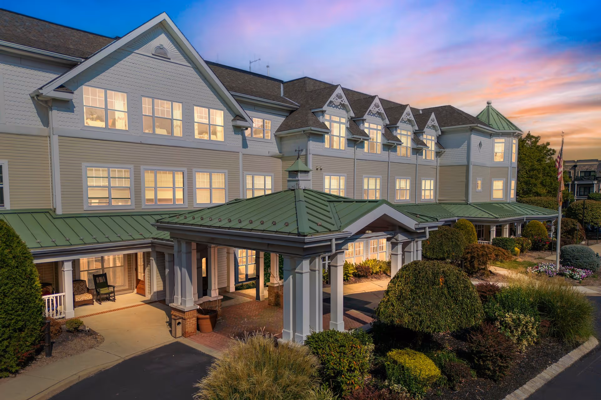 Exterior view of a large senior living facility building at sunset with multiple windows lit from inside, a green metal roof, a covered entrance with white columns, and well-maintained landscaping including bushes and flowers.