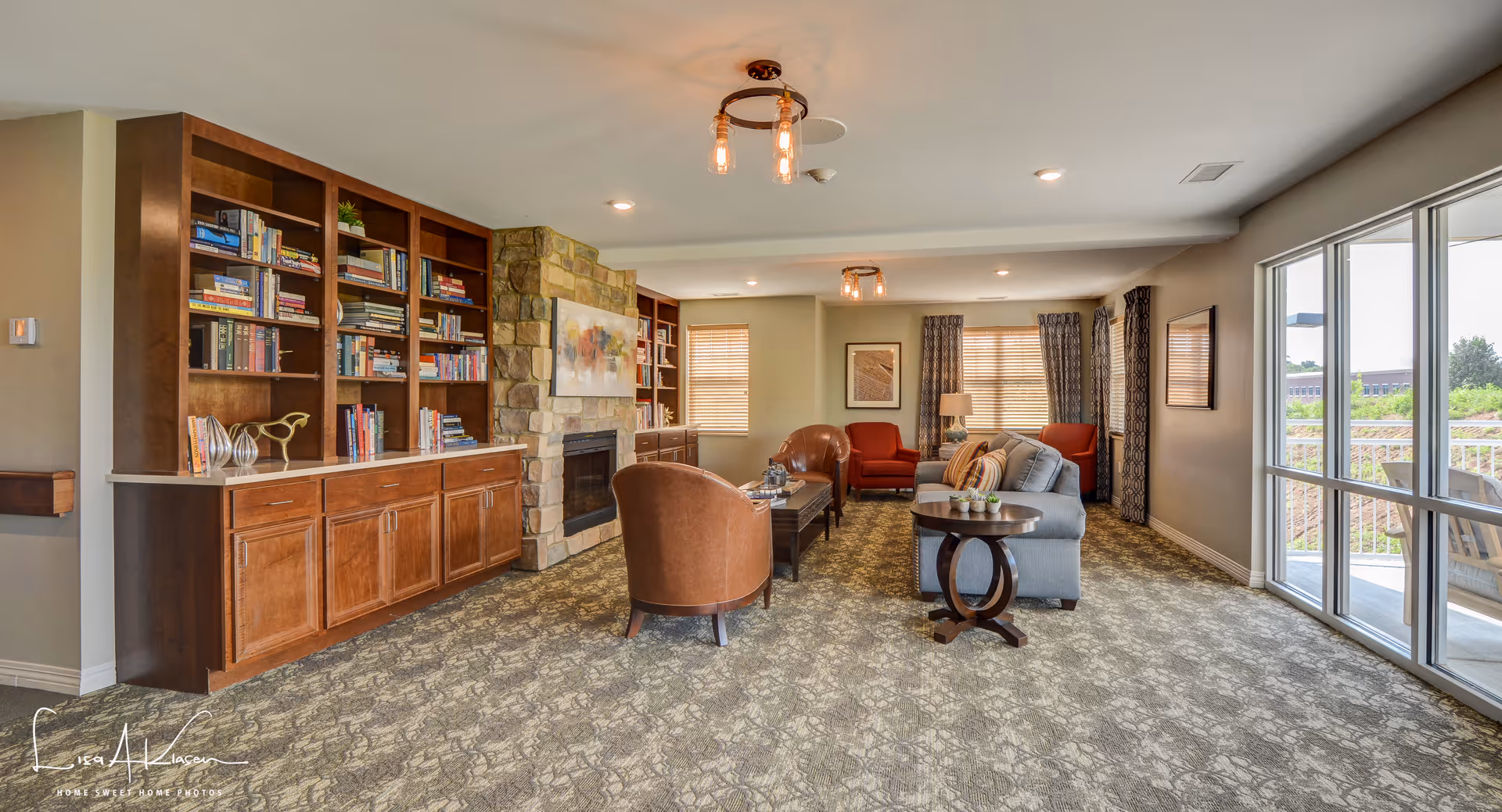 A cozy senior living facility common area with a stone fireplace, built-in wooden bookshelves filled with books, comfortable seating including leather chairs and a gray sofa, carpeted floor, large windows with curtains letting in natural light, and a small round wooden table with decorative items.