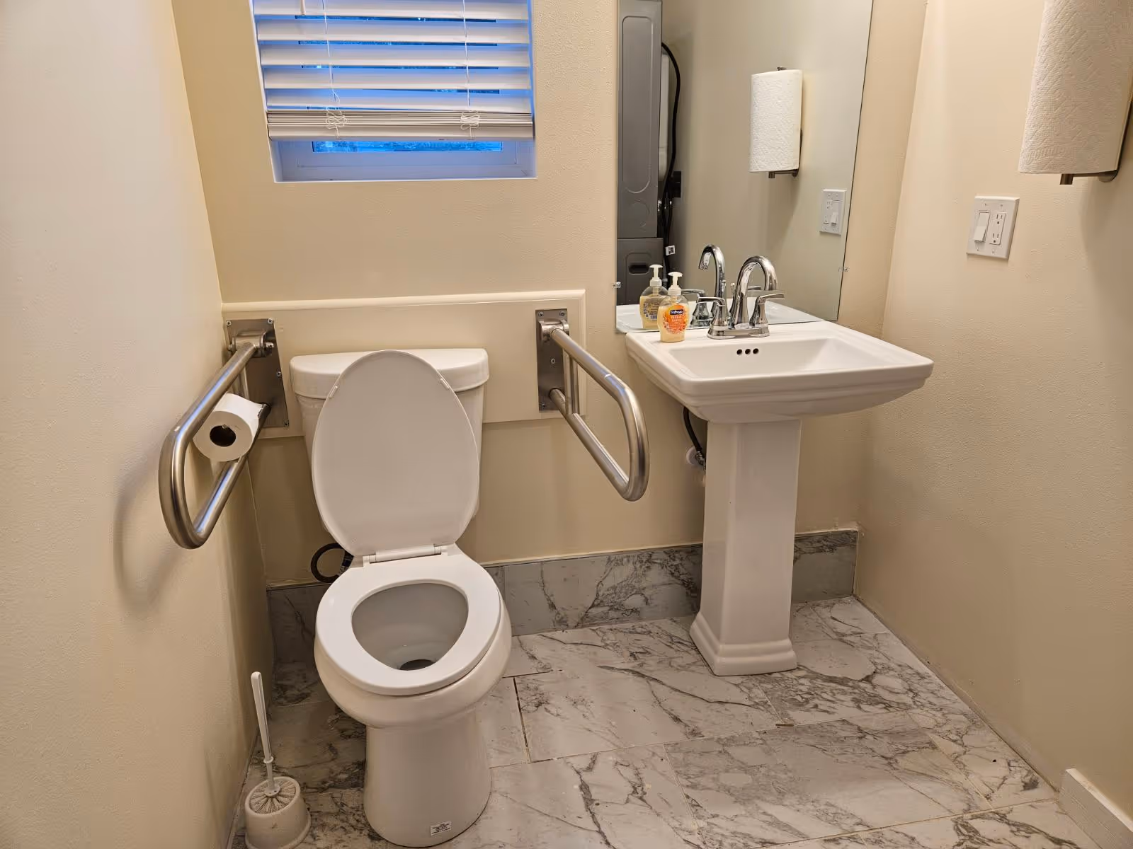 Accessible bathroom with a toilet flanked by stainless steel grab bars, a pedestal sink with soap dispensers, and a small window.