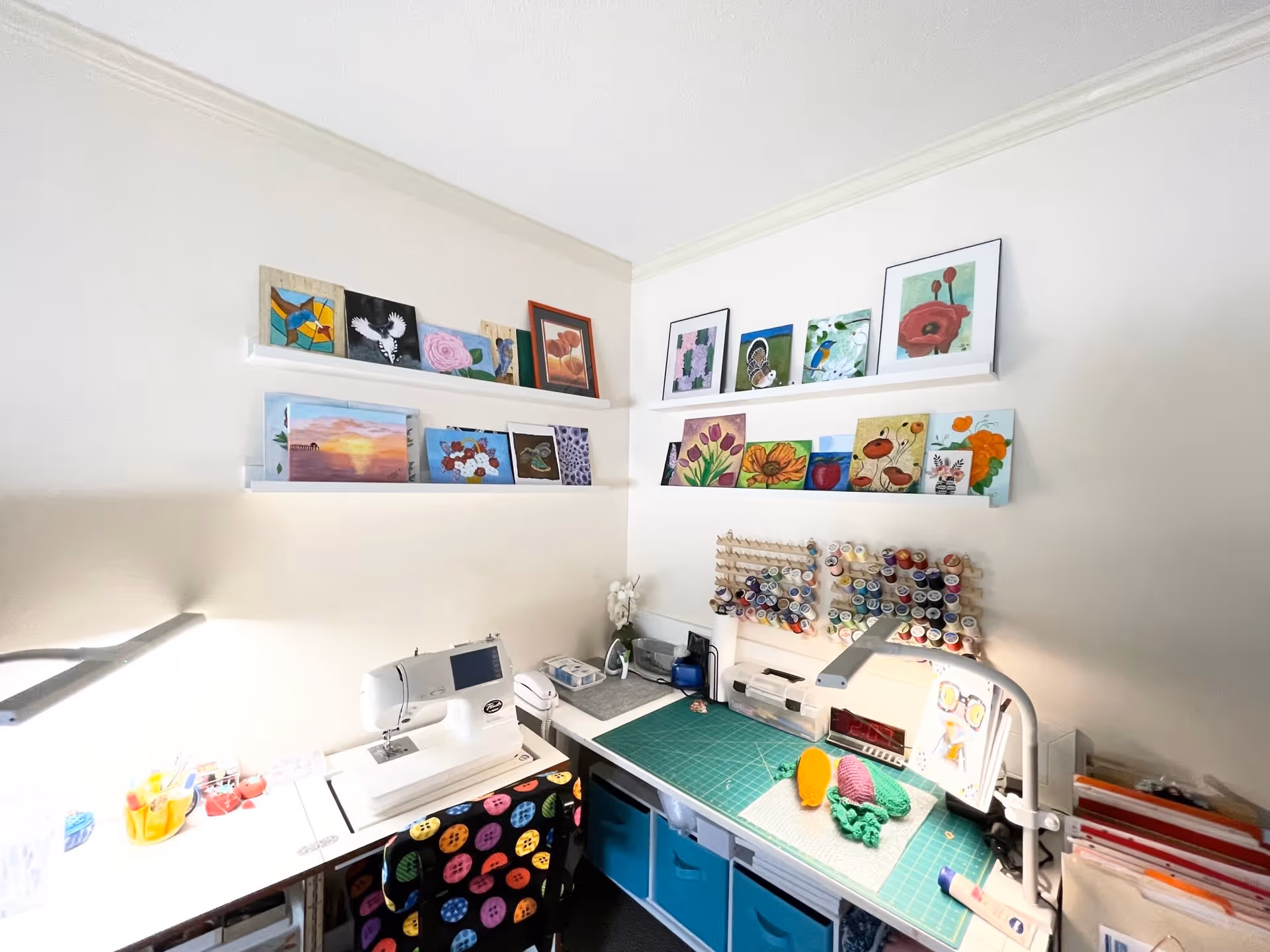 A bright craft room corner with a sewing machine on a white table, colorful fabric with button patterns on a chair, and a green cutting mat on another table. The walls have shelves displaying various small paintings of flowers, birds, and other nature scenes. There are organized spools of thread on racks above the cutting mat, and various craft supplies are neatly arranged on the tables.