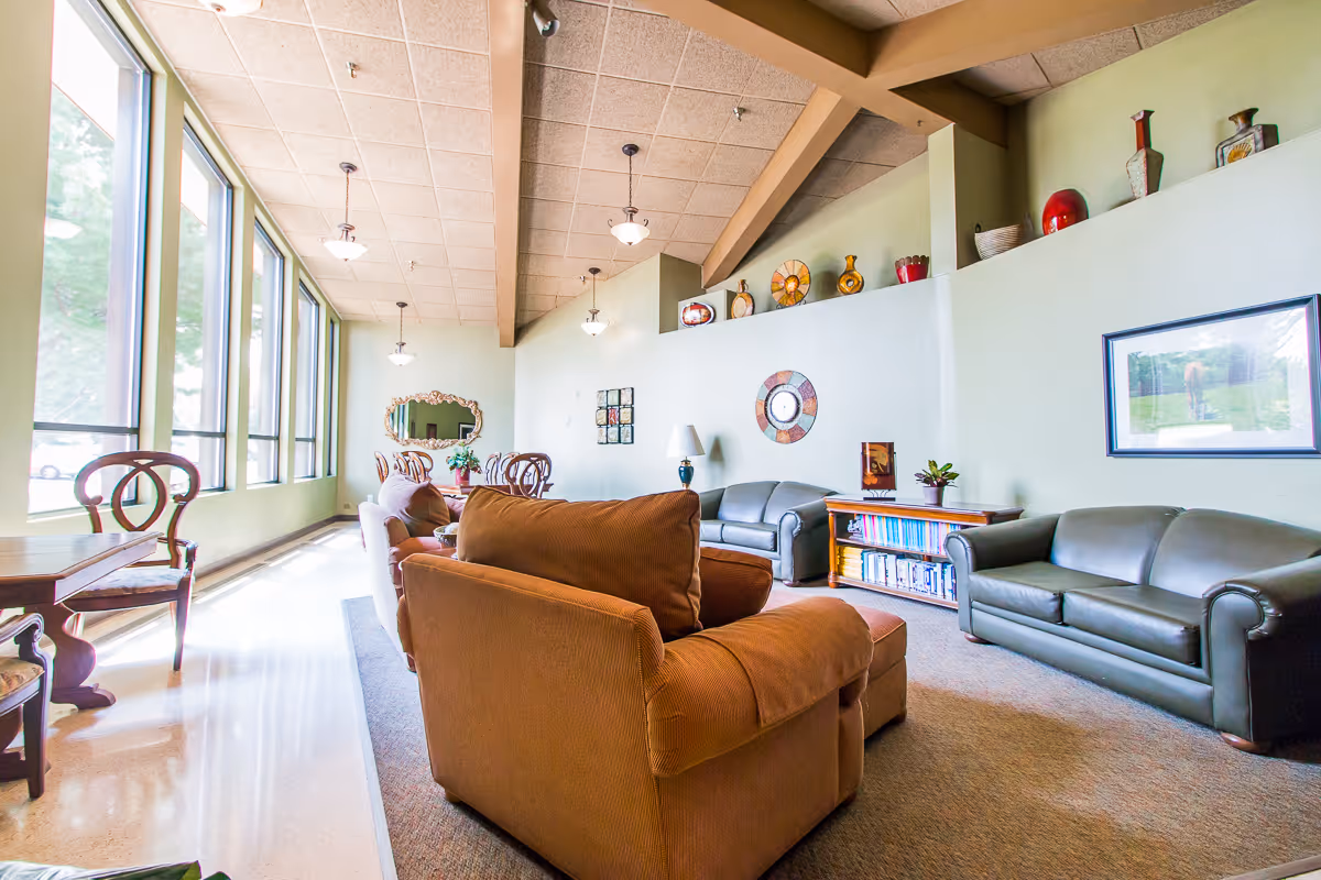 A bright and spacious living room area with large windows letting in natural light. The room features comfortable seating including a brown armchair and two dark green sofas. There is a wooden bookshelf filled with books, decorative vases on a high ledge, a framed picture on the wall, and a table with chairs near the windows.