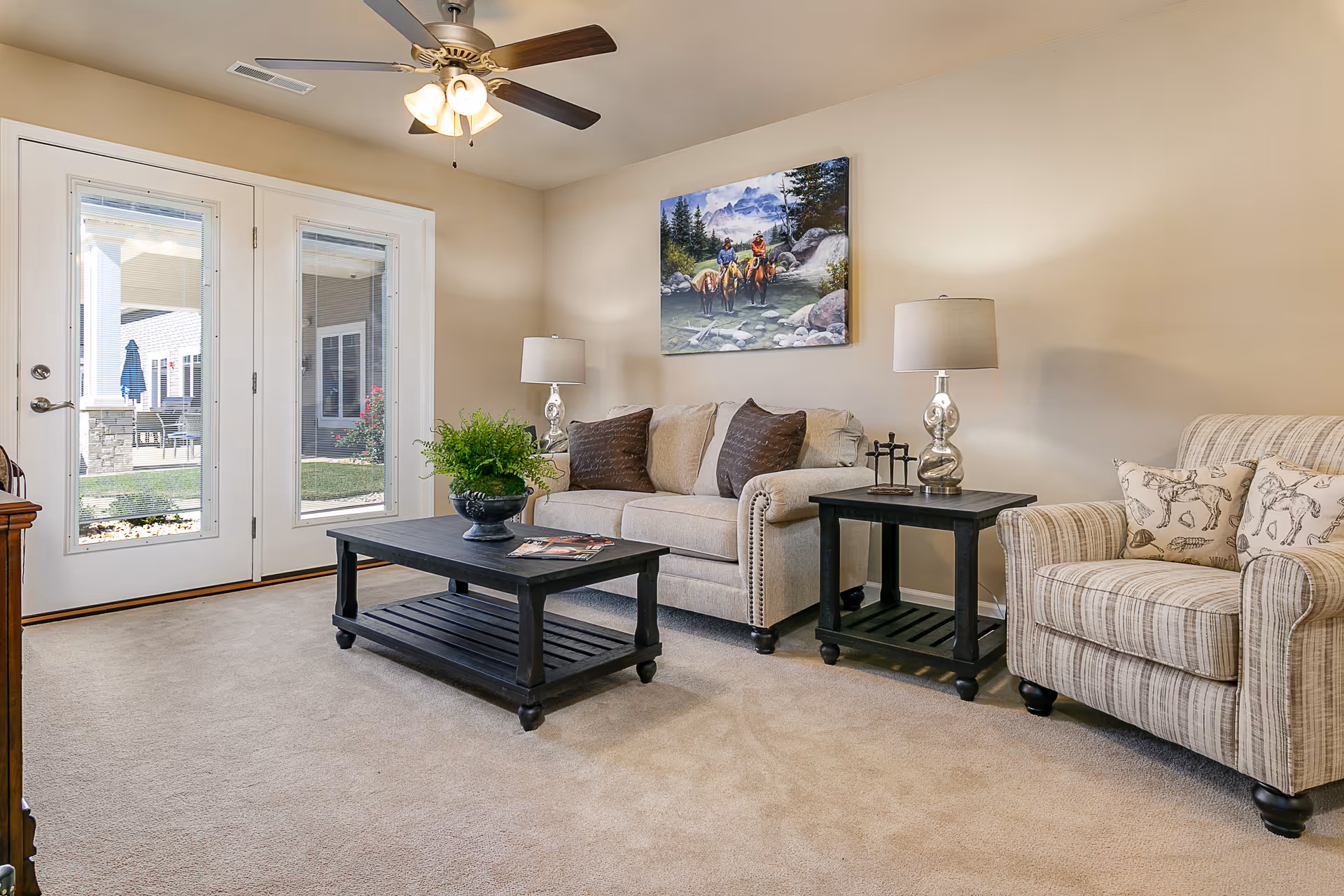 A cozy living room with beige walls and carpet, featuring a beige sofa with brown pillows, a striped armchair with horse-patterned cushions, a black coffee table with a plant and magazines, and a matching black side table with a lamp. A ceiling fan with lights is mounted on the ceiling, and a painting of two people riding horses in a mountainous landscape hangs on the wall. French doors with glass panes open to an outdoor patio area.