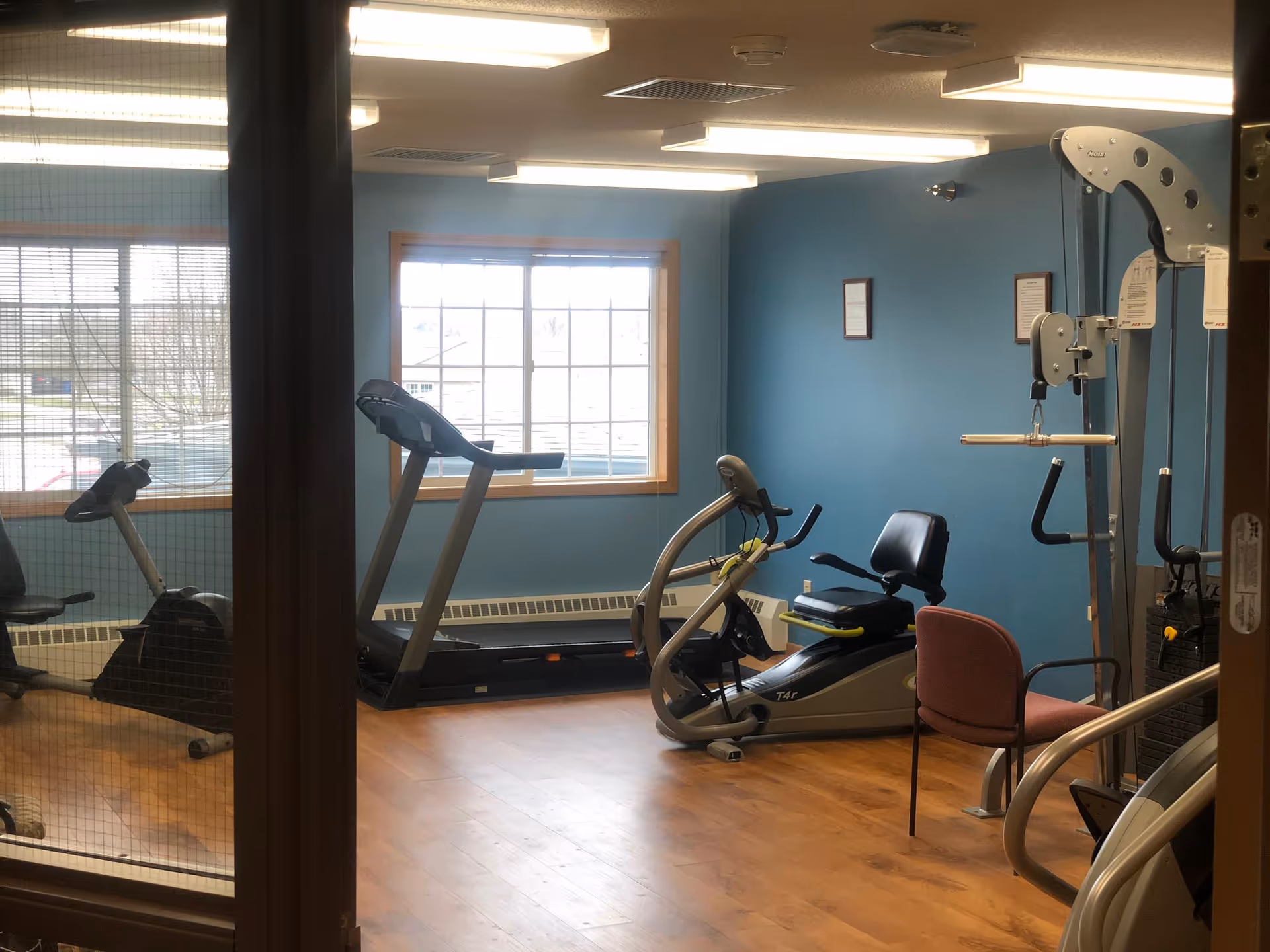 A small fitness room with exercise equipment including a treadmill, a recumbent bike, and a multi-function weight machine. The room has blue walls, a large window letting in natural light, and wood flooring. There is also a red chair placed near the weight machine.