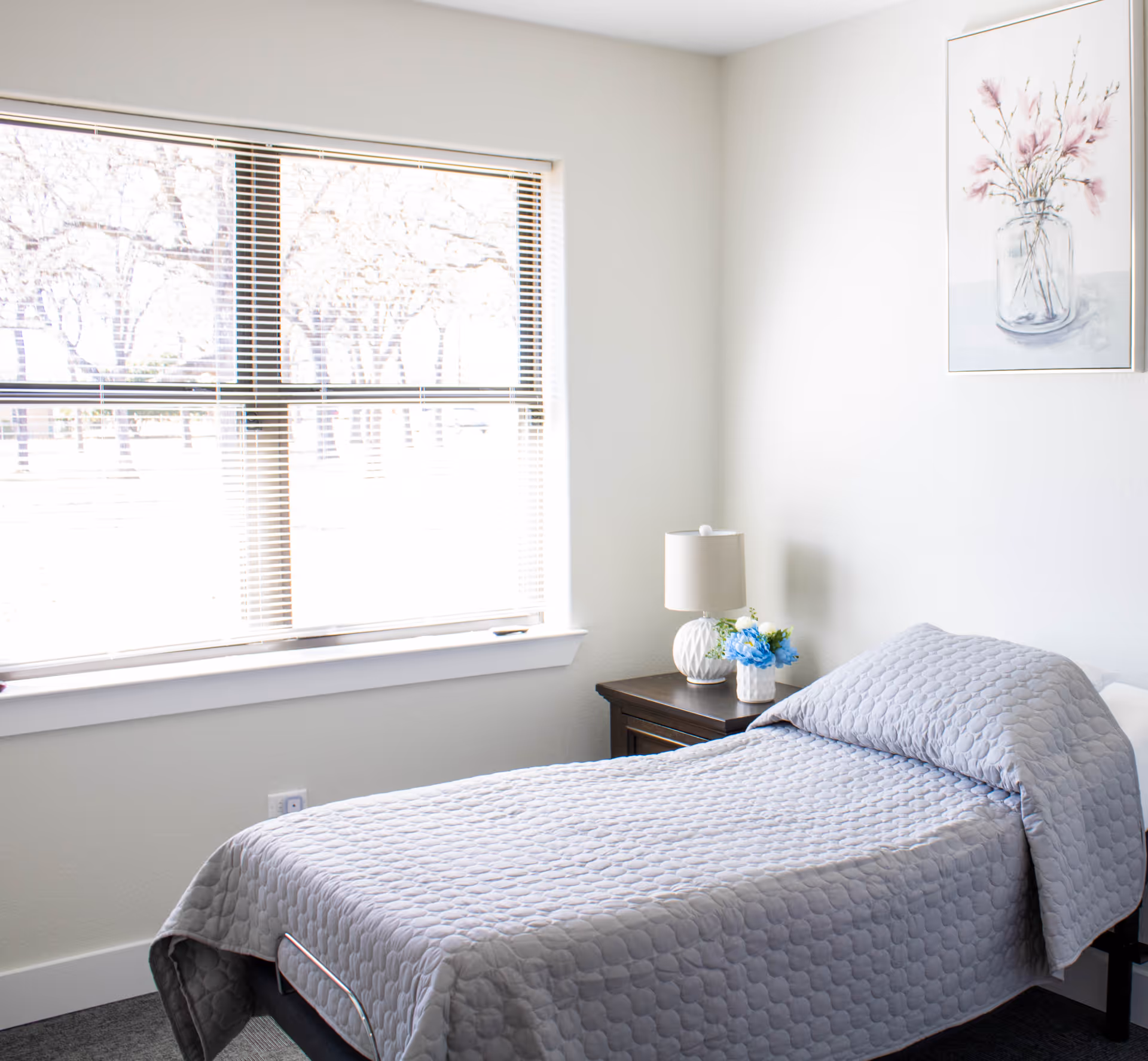 A neatly made single bed with a gray quilted bedspread in a bright room with a large window covered by blinds. Next to the bed is a dark wooden nightstand with a white lamp and a small vase of blue and white flowers. A framed picture of flowers in a vase hangs on the white wall above the nightstand.