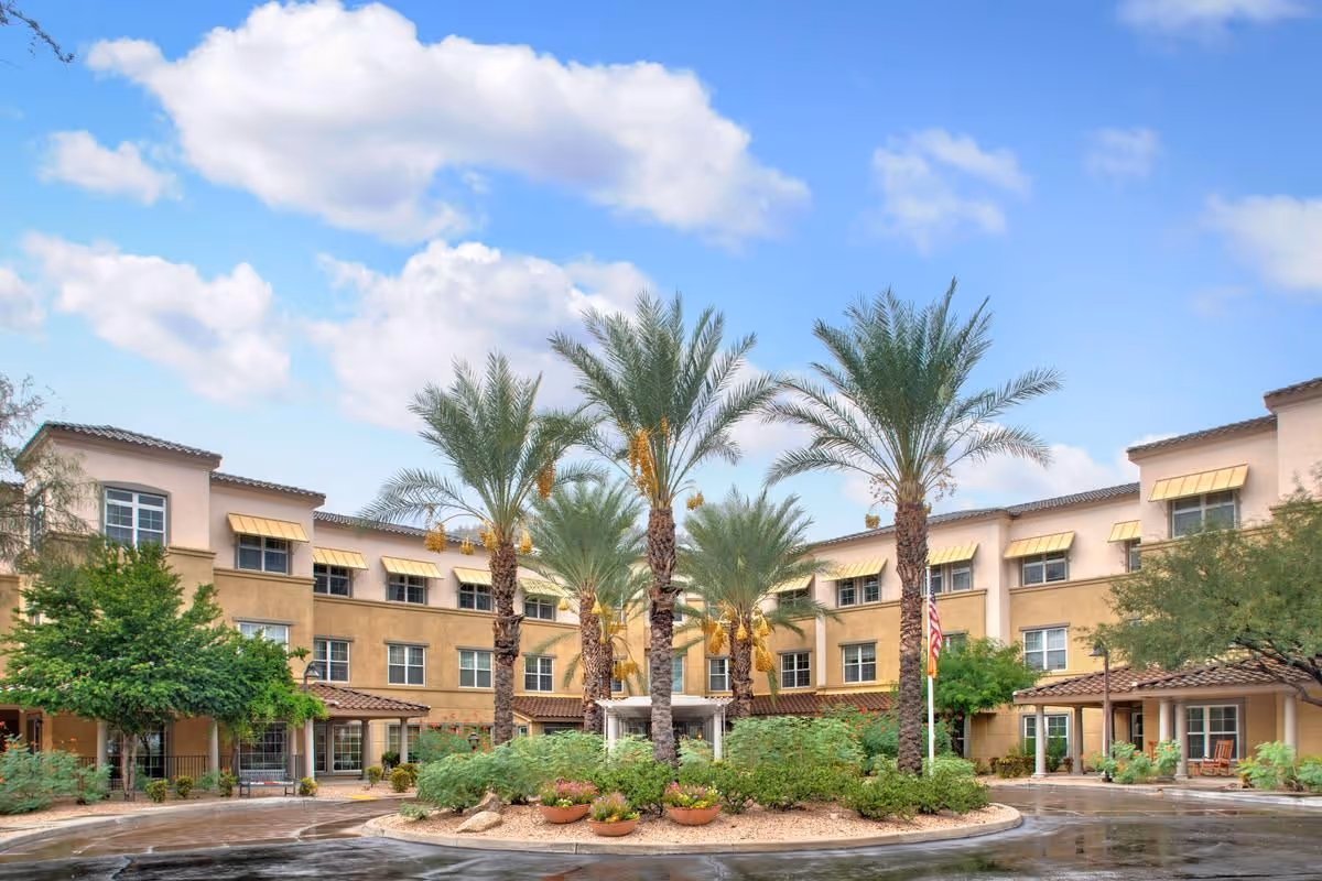 Exterior view of a three-story senior living facility building with beige and light brown walls, multiple windows with yellow awnings, and a circular driveway with landscaped greenery and tall palm trees in the center under a partly cloudy blue sky.