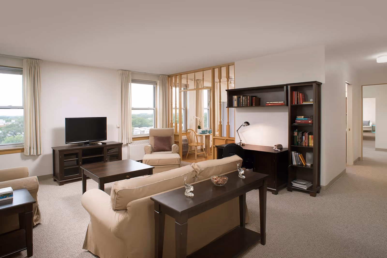 A bright and spacious living room in a retirement community featuring beige sofas, a wooden coffee table, a TV on a wooden stand, a comfortable armchair, and a wooden desk with a lamp and bookshelf filled with books. Large windows with light curtains allow natural light to fill the room. A small dining area with a wooden table and chairs is visible in the background, separated by wooden spindles. A hallway leads to other rooms.
