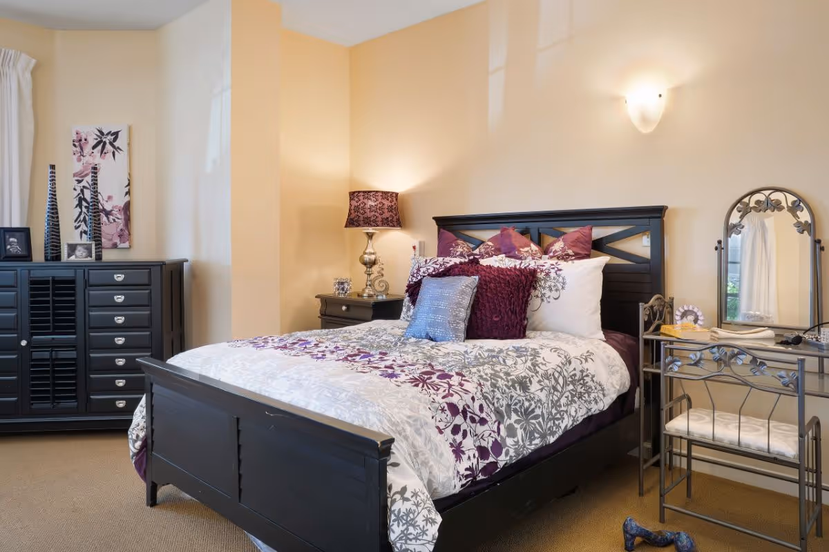 Well-decorated bedroom with a dark wooden bed dressed in patterned bedding and multiple pillows, a dresser, bedside tables, and a small vanity.