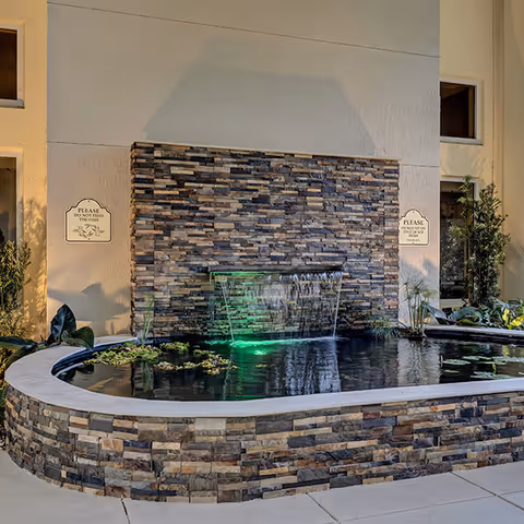 A stone water feature with a cascading waterfall and green-lit pond surrounded by plants against a building wall.