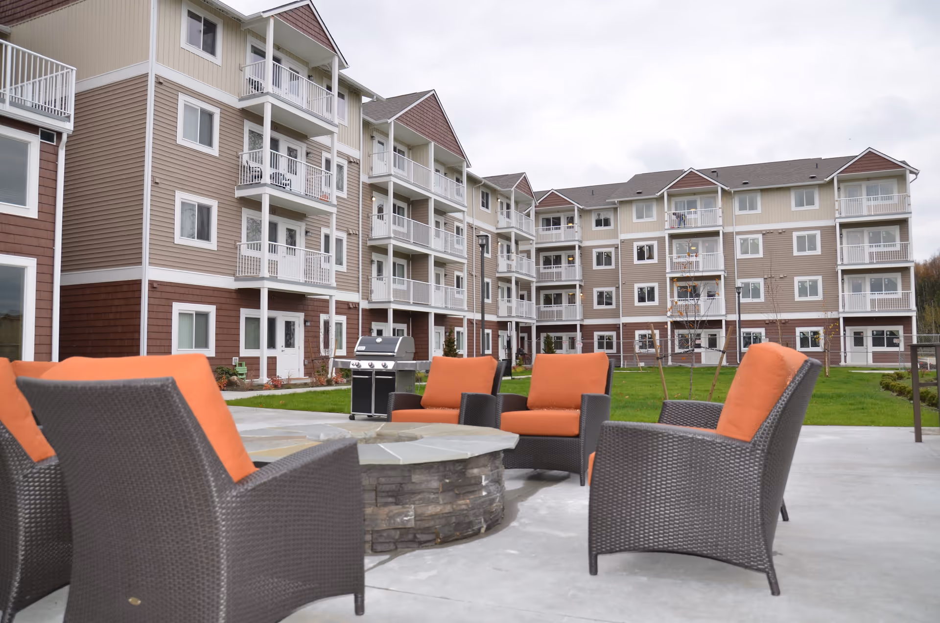 Outdoor seating area with four wicker chairs featuring orange cushions arranged around a stone fire pit, situated on a concrete patio in front of a multi-story residential building with balconies.