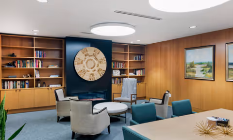 A cozy communal lounge with bookshelves, armchairs, a round table and wood-paneled walls under circular ceiling lights.