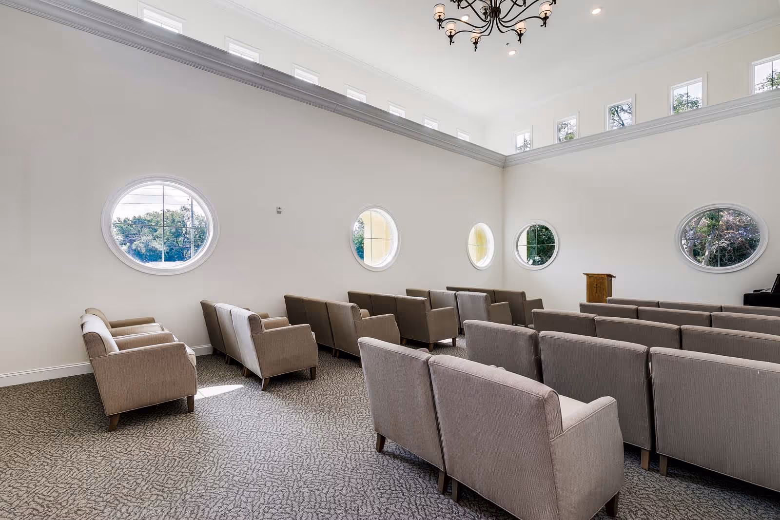 A bright room with high ceilings and multiple round windows along the walls. The room contains several rows of beige upholstered chairs arranged facing a wooden podium and a piano in the corner. A decorative chandelier hangs from the ceiling.