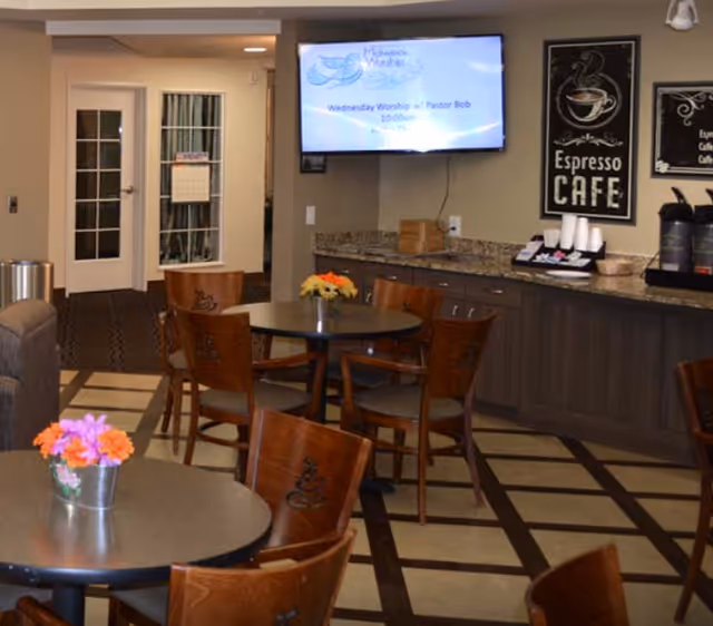 A small cafe-style dining area with round tables and wooden chairs, a countertop espresso station, and a wall-mounted TV.
