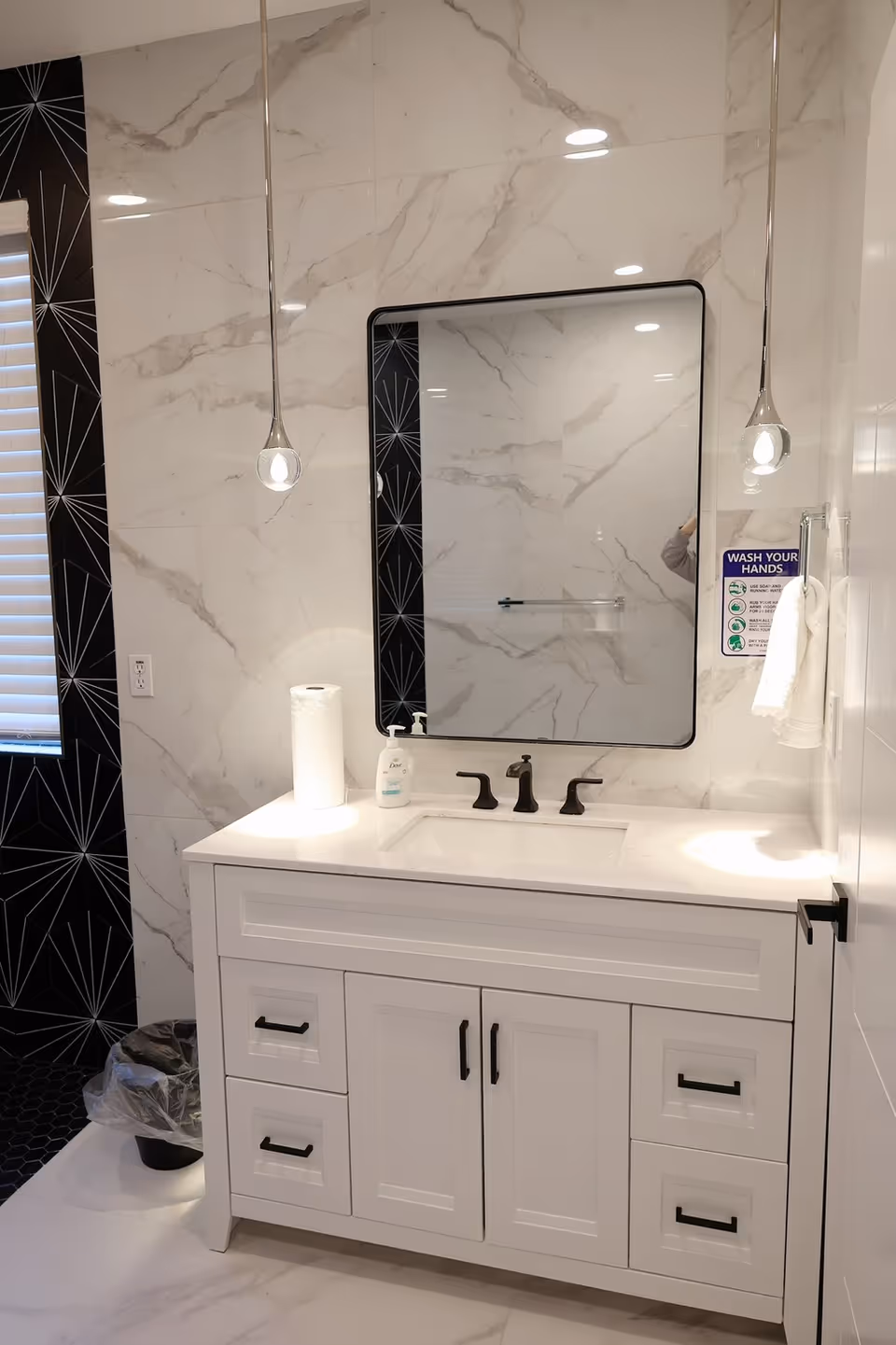 A modern bathroom vanity with a white cabinet and black handles, a rectangular sink with black faucet fixtures, a large rectangular mirror with a black frame, and two pendant lights hanging from the ceiling. The walls are covered in white marble tiles with gray veining, and there is a black patterned tile accent on one side. A paper towel roll and a bottle of hand soap are on the countertop. A small sign on the wall next to a white hand towel reads 'WASH YOUR HANDS'. A trash can with a plastic liner is visible on the floor near the window with white blinds.