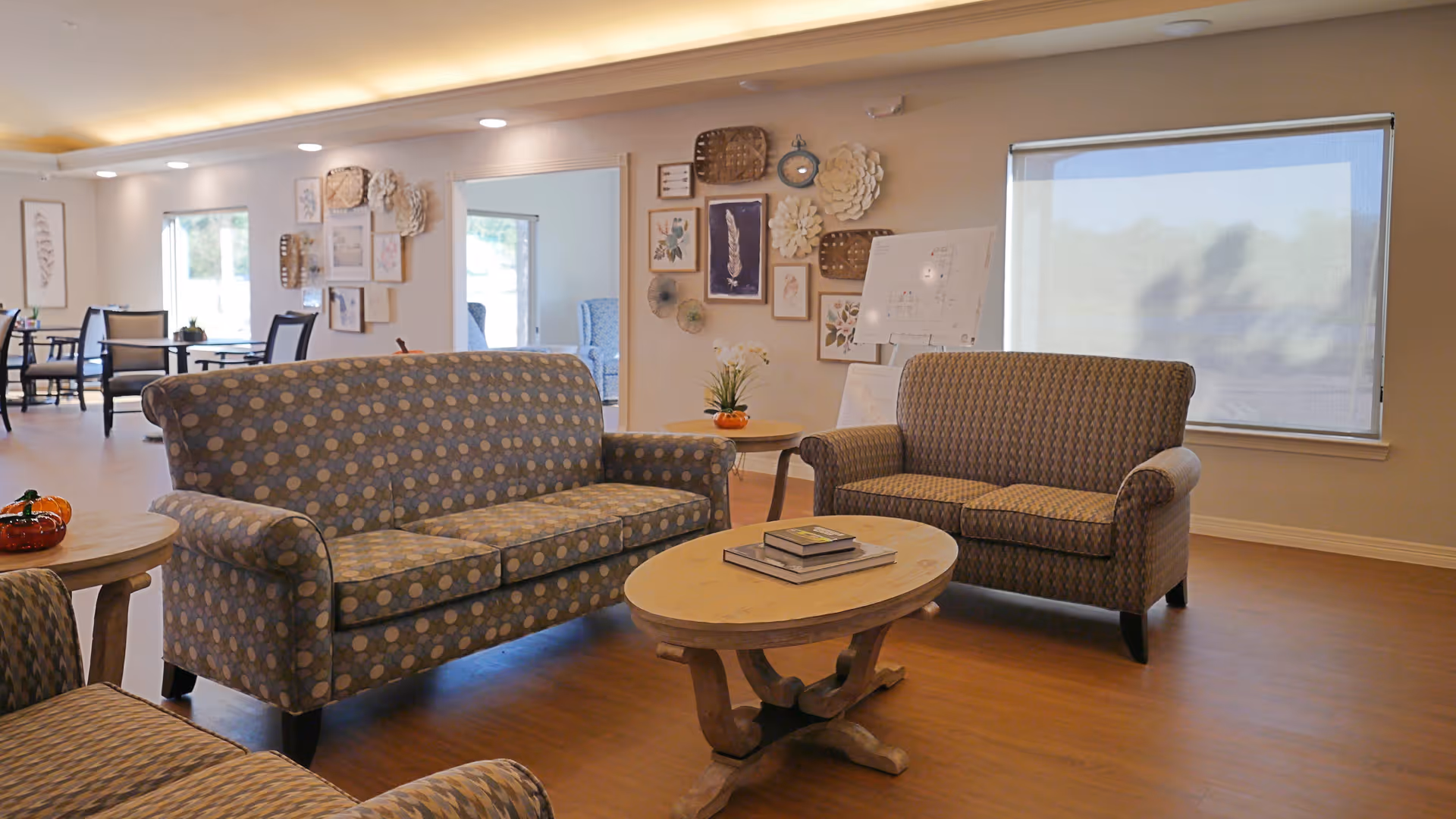 A cozy living room area in Arbor House Fredericksburg featuring two patterned sofas and a matching armchair arranged around a wooden coffee table with books on it. The room has wooden flooring, a large window with a roller shade, and a wall decorated with framed artwork and decorative items. In the background, there are dining tables and chairs near windows letting in natural light.