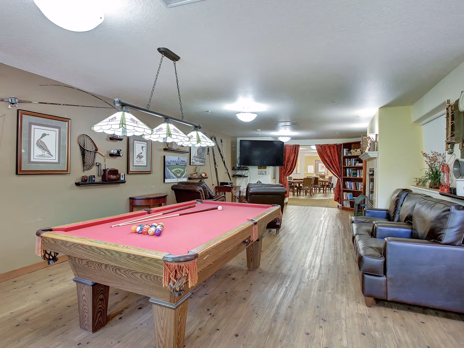 A recreational room in a senior living facility featuring a red felt pool table with pool cues and balls arranged for a game. The room has wooden flooring, leather sofas, a wall-mounted TV, and various wall decorations including framed pictures and fishing equipment. In the background, there is a dining area visible through an open doorway with red curtains.