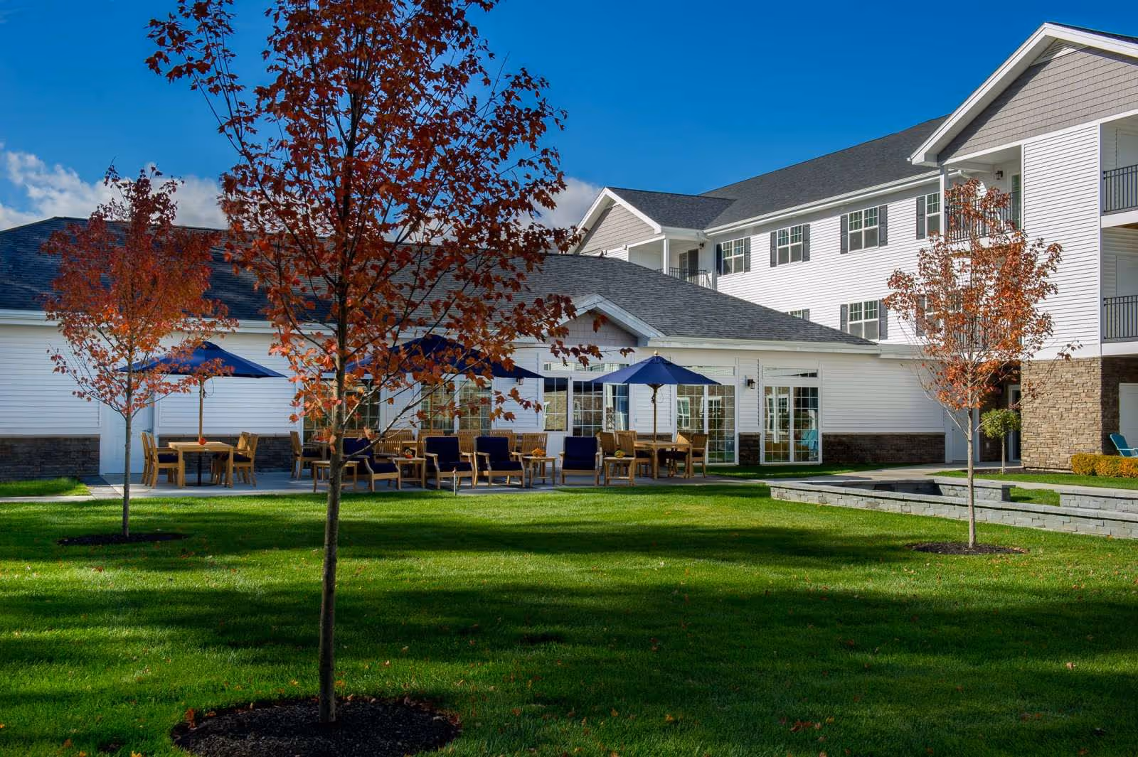 Green courtyard with young trees, outdoor seating and umbrellas in front of a multi-story white senior living building.