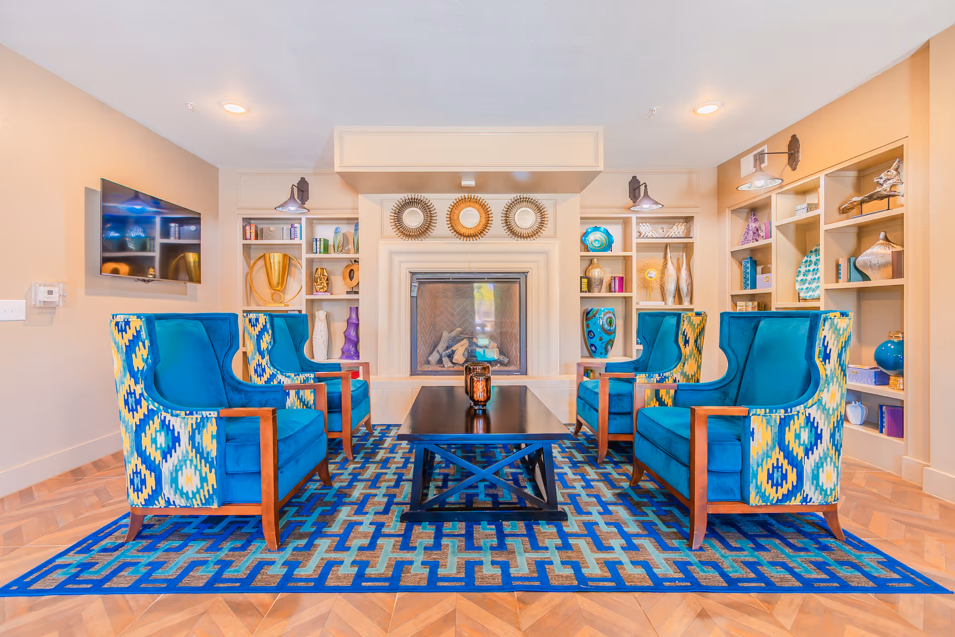 A bright and colorful living room area with four blue and patterned armchairs arranged around a dark wooden coffee table on a blue geometric patterned rug. The room features a fireplace with decorative mirrors above it and built-in shelves on either side filled with various decorative items. A flat-screen TV is mounted on the left wall.