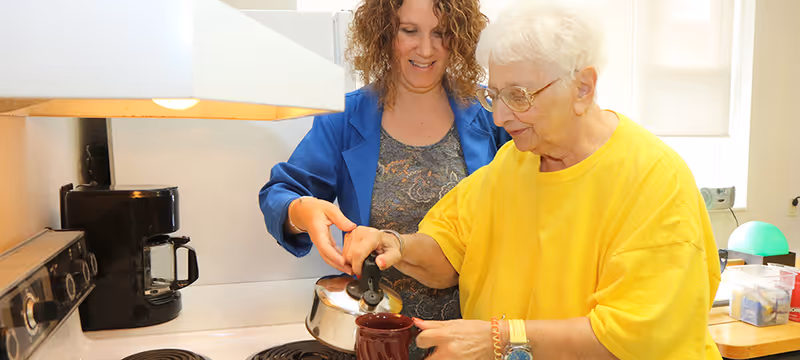 An elderly woman in a yellow shirt is pouring a beverage from a kettle into a mug with the assistance of a younger woman in a blue jacket in a kitchen setting.