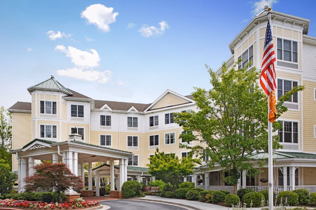 Exterior view of a multi-story senior living facility building with cream and yellow siding, multiple windows, a covered entrance, landscaped bushes and trees, an American flag and an orange flag on a flagpole, under a partly cloudy blue sky.