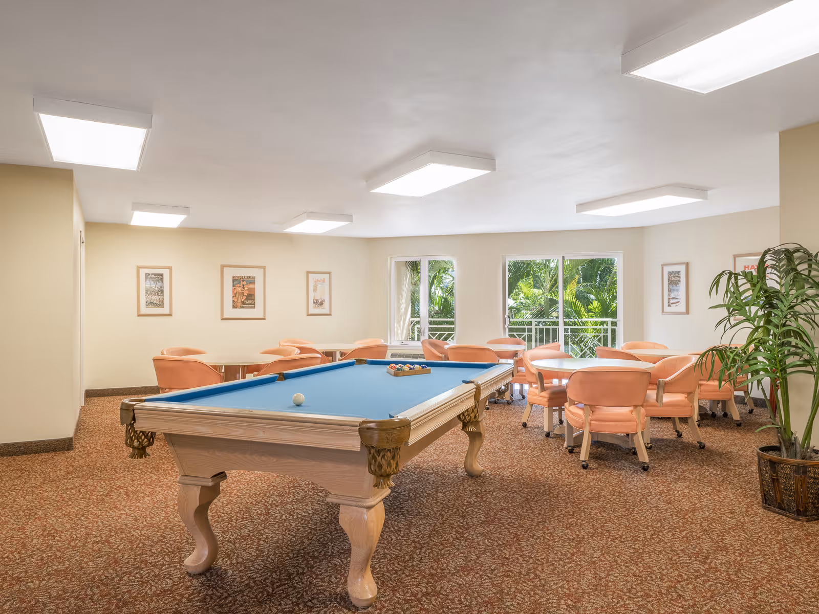A bright recreational room with a pool table in the center and several round tables surrounded by peach-colored chairs. The room has large windows showing green foliage outside, beige walls with framed pictures, and a patterned carpet floor. A potted plant is visible on the right side.