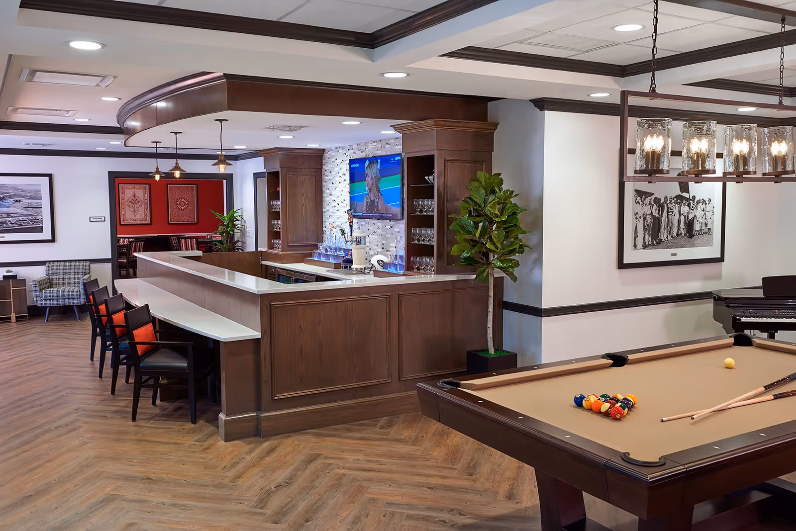 A recreational room featuring a pool table with billiard balls and cues, a wooden bar with stools, a flat-screen TV mounted on a tiled wall behind the bar, and a potted plant. The room has wood flooring, framed artwork on the walls, and modern ceiling lights.