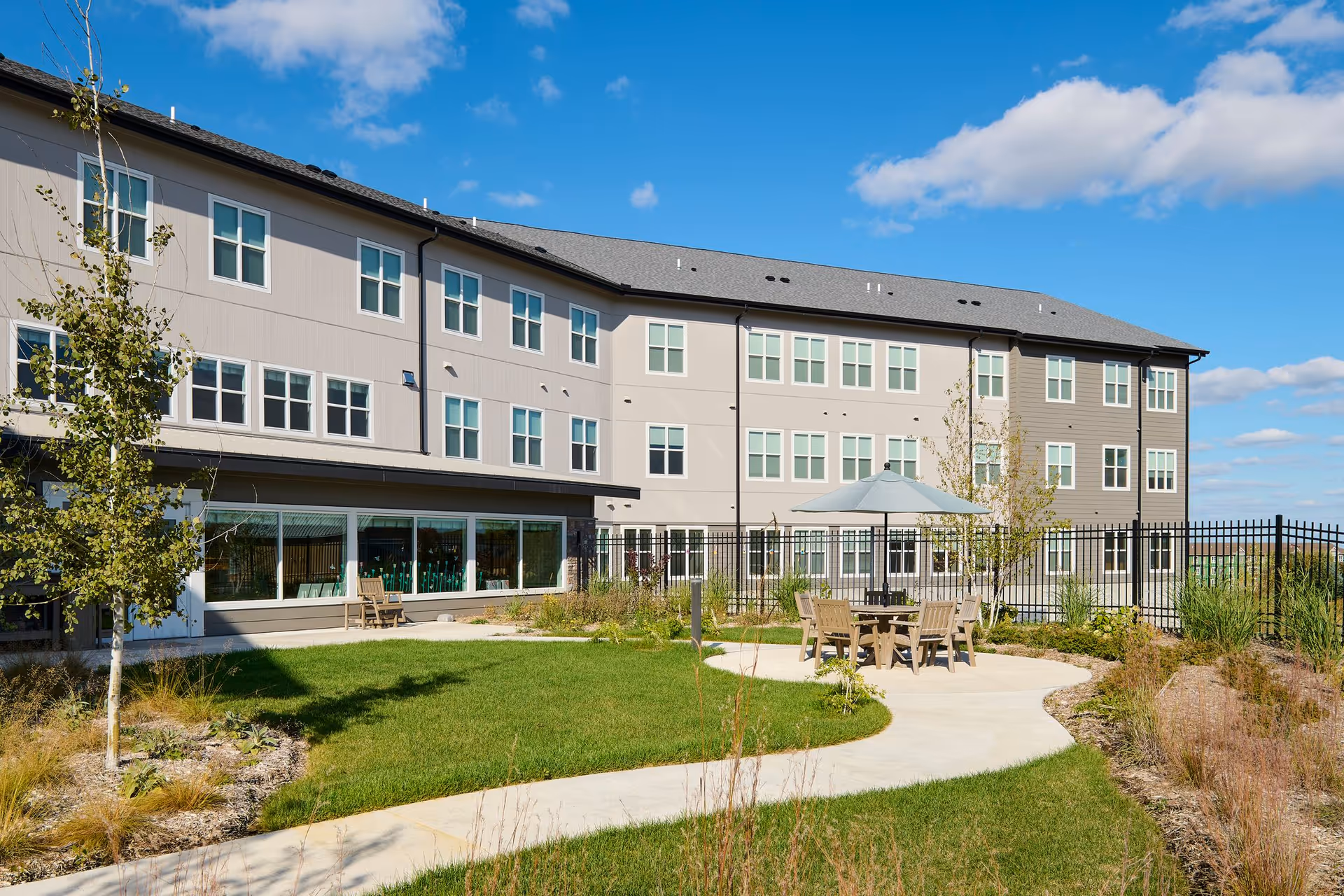 Courtyard with a round table, chairs and umbrella beside a three-story senior living building under a blue sky.