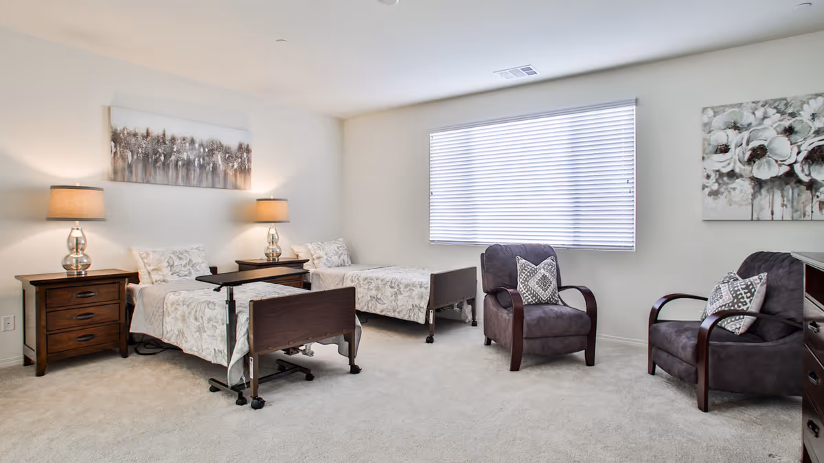 A bright and clean assisted living bedroom with two single beds, each with floral bedding and a bedside table with a lamp. There are two cushioned armchairs with patterned pillows near a large window with blinds. The walls are decorated with two paintings featuring neutral tones.