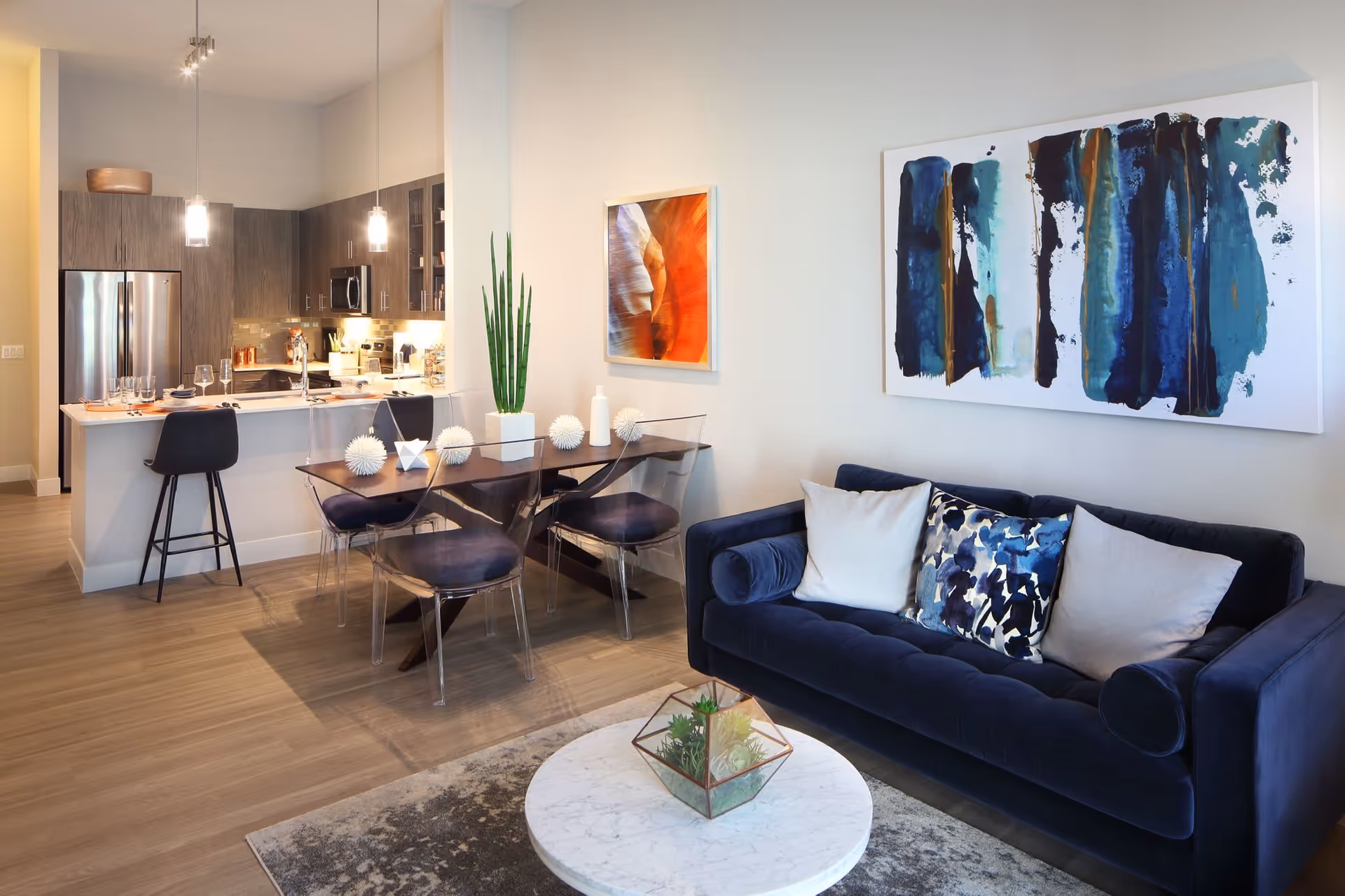 Modern open-concept living space featuring a dark blue velvet sofa with decorative pillows, a round marble coffee table with a geometric terrarium centerpiece, a wooden dining table with transparent chairs, and a kitchen area with dark wood cabinets, stainless steel appliances, and pendant lighting.