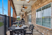 Outdoor covered patio area with black metal tables and chairs, ceiling fans, and stone wall exterior with windows. The patio overlooks a grassy area and is enclosed by a black metal railing.