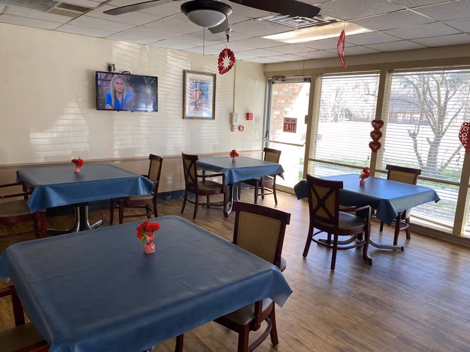 A dining room with several tables covered in blue tablecloths, each with a small red flower centerpiece. There are wooden chairs around the tables. A television is mounted on the wall showing a woman in a blue top. Large windows with blinds let in natural light, and there are red heart decorations hanging from the ceiling and on the window. A door with a 'Not an Exit' sign is visible.