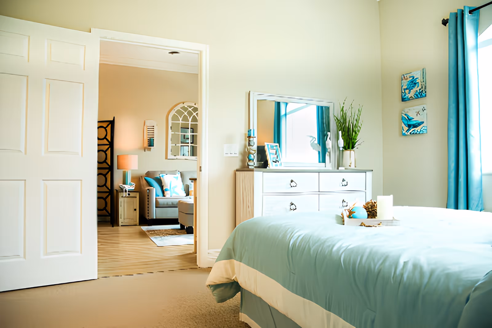 A bright bedroom with a light blue bedspread and a white dresser with a mirror. There are decorative items on the dresser including a plant and framed photos. The room has light-colored walls and a window with blue curtains. Through an open door, a living room area with a gray armchair, side table, lamp, and wall mirror is visible.