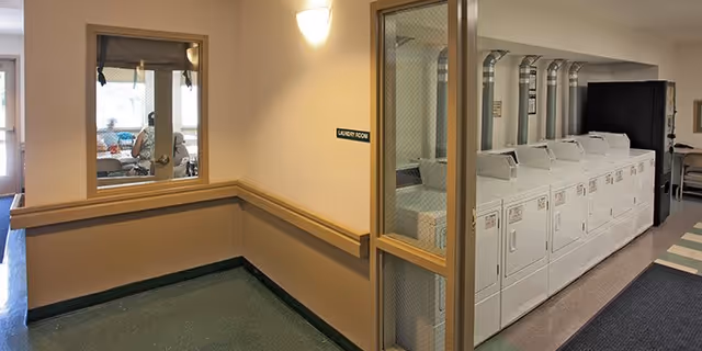 Interior view of a laundry room in a senior living facility with a row of white washing machines and dryers along the right wall. There is a window and a door labeled 'Laundry Room' on the left side, and a person can be seen sitting in a room through the window. The walls are beige with a wooden chair rail, and the floor has green and gray carpeting.