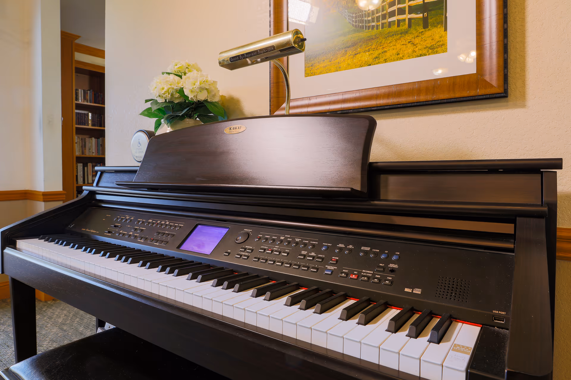 A digital piano keyboard with control buttons and a small screen, placed against a wall with a framed picture above it and a vase of white flowers on top. A reading lamp is attached to the piano. A bookshelf is visible in the background.