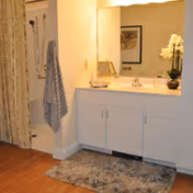 Bathroom area with a white vanity cabinet, a large mirror above the sink, a decorative orchid plant on the countertop, a framed picture reflected in the mirror, a striped towel hanging near a shower with a curtain, and a textured rug on the wooden floor.