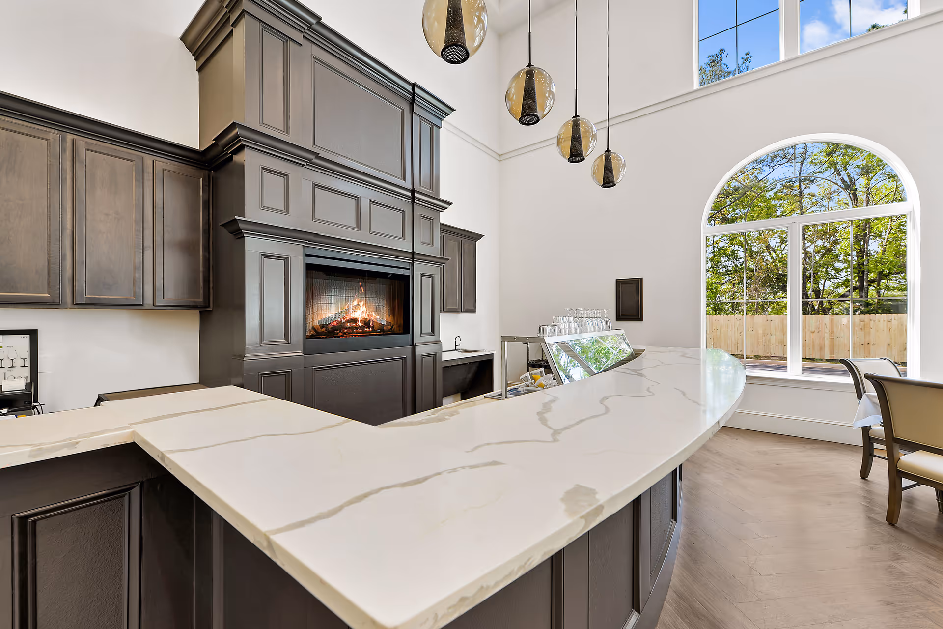 Interior view of a modern dining area with a large curved marble countertop bar, dark wood cabinetry, a built-in fireplace, and three hanging pendant lights. There is a large arched window showing trees and a wooden fence outside, with chairs and a table partially visible to the right.
