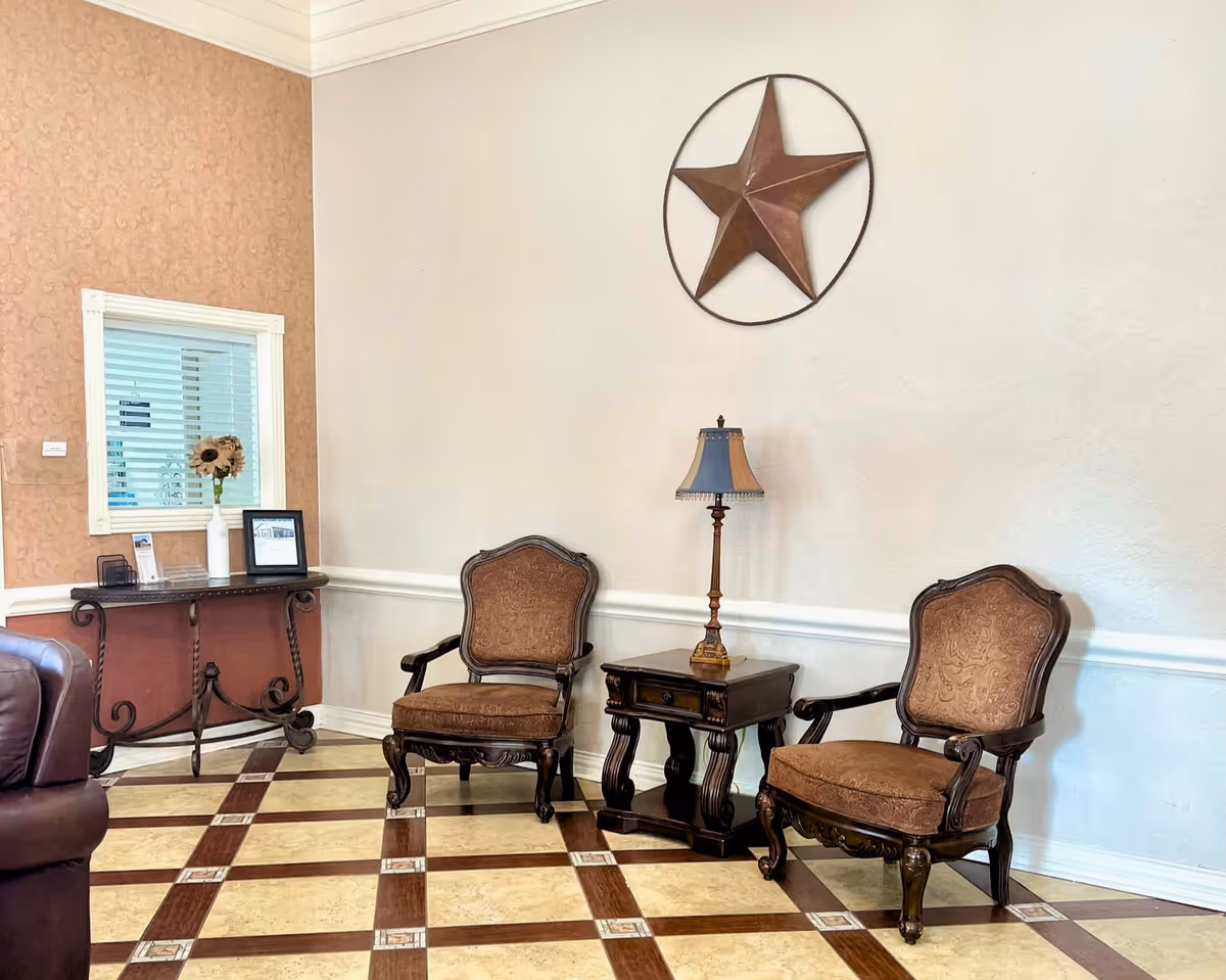 A sitting area with two ornate brown upholstered armchairs and a dark wooden side table with a decorative lamp on it. The floor has a checkered pattern with beige and brown tiles. On the wall above the chairs is a large metal star decoration. To the left, there is a small table with a vase of sunflowers and some framed items, next to a window with blinds.