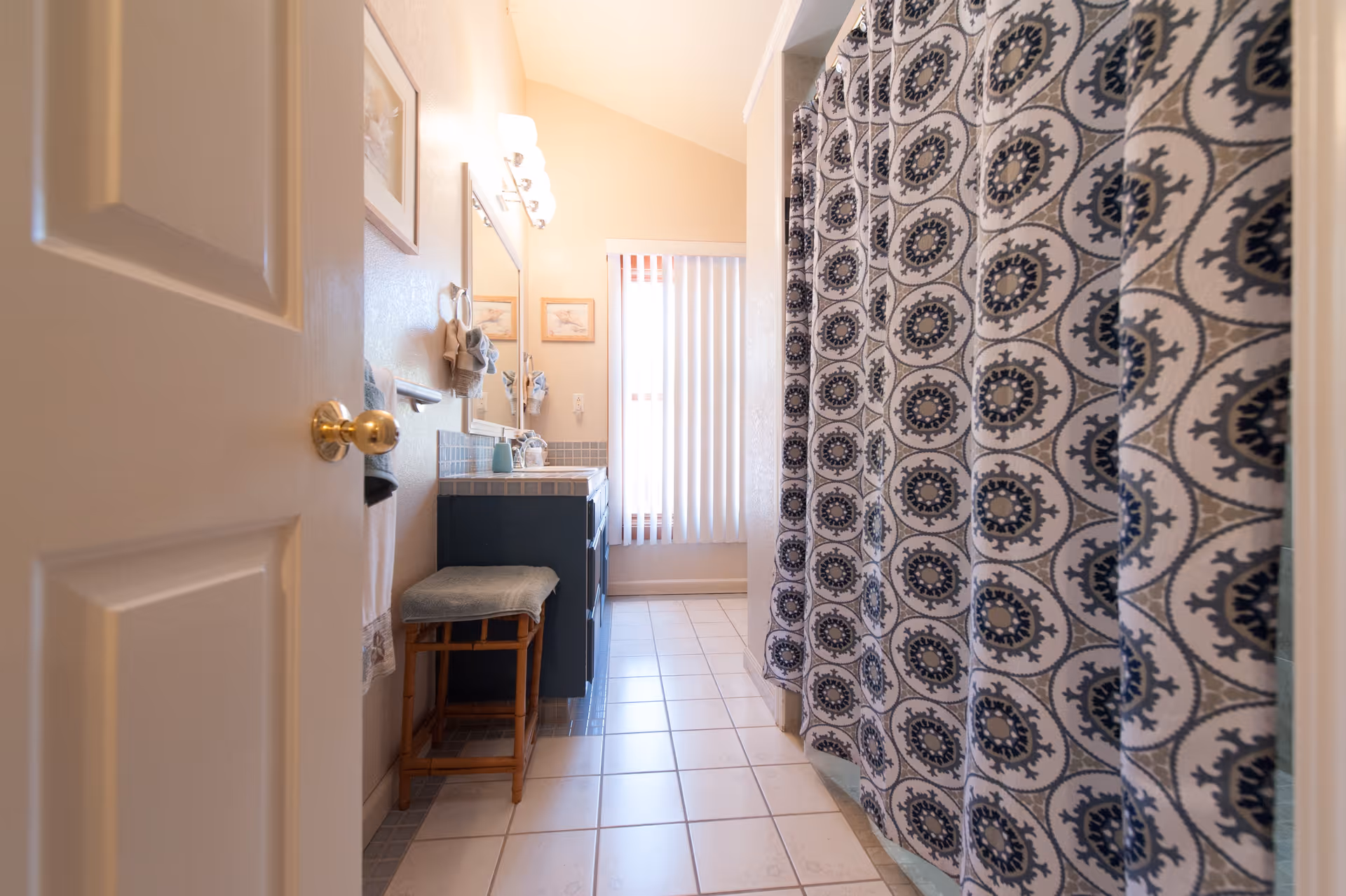 View into a bathroom with a patterned shower curtain on the right, a vanity with a mirror and lights on the left, a cushioned stool under the counter, and a window with vertical blinds at the far end.