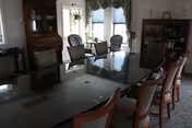 A formal dining room with a large rectangular table surrounded by multiple wooden chairs with cushioned seats. The room features windows with blue curtains, a wooden cabinet, and a bookshelf. The lighting is natural from the windows, and the overall decor is traditional.