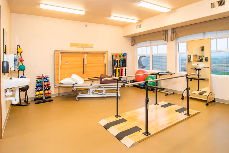 A bright physical therapy room with parallel bars on a wooden platform for walking exercises, a treatment table with pillows, various colorful exercise balls and weights, a large mirror, and a wall-mounted sink. The room has large windows with a view outside and beige flooring.