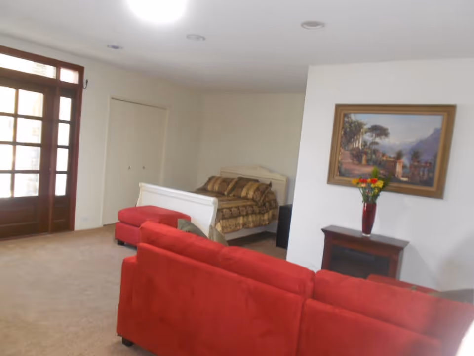 A cozy room with a red sofa and matching ottoman, a bed with patterned bedding, a wooden door with glass panels, a small table with a vase of flowers, and a framed painting on the wall.