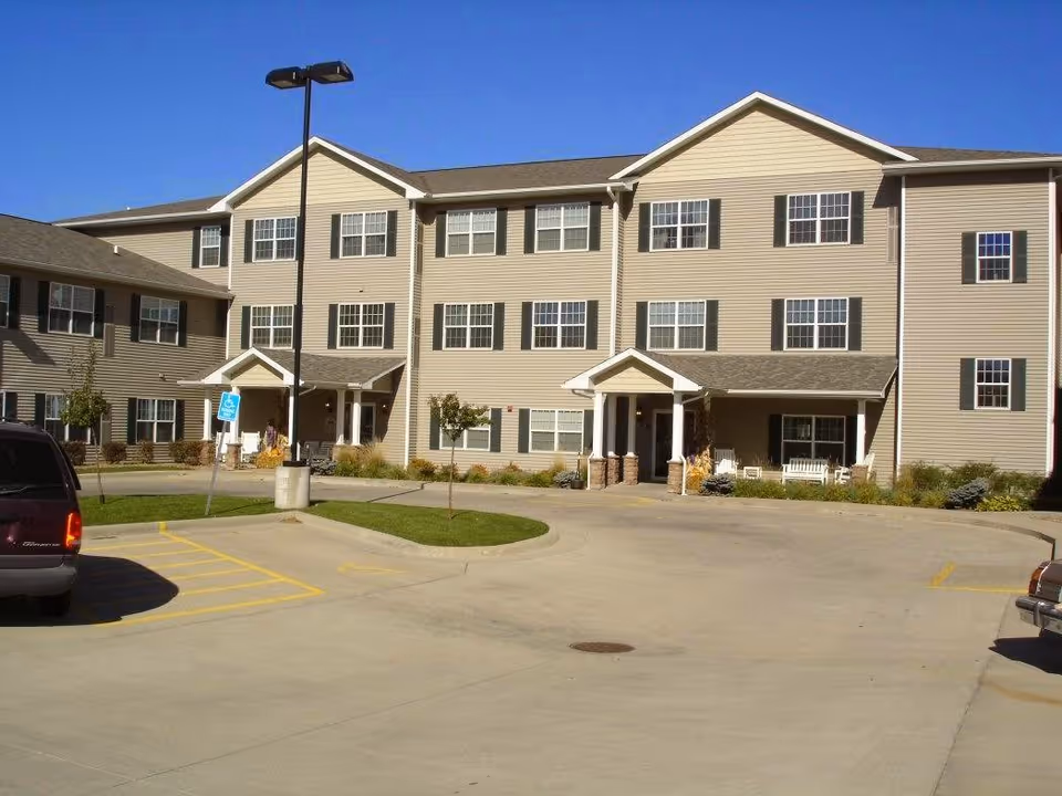 Exterior view of a three-story beige senior living facility building with multiple windows and two covered entrances. There is a parking lot in front with a handicapped parking sign and a few vehicles parked. The sky is clear and blue.